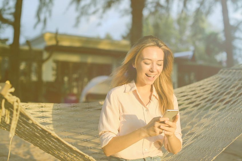 Young happy woman using smartphone and sitting in wicker hammock on tropical resort.
